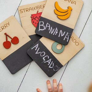 May include: Wooden learning cards with chalkboard surfaces featuring the words "banana", "strawberry", "cherry", and "avocado". The cards have illustrations of the fruits on the front and a chalkboard surface on the back.