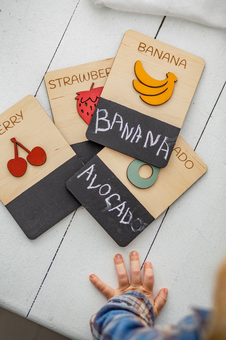 May include: Wooden learning cards with chalkboard surfaces featuring the words "banana", "strawberry", "cherry", and "avocado". The cards have illustrations of the fruits on the front and a chalkboard surface on the back.