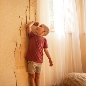 May include: A young child stands next to a wooden castle-shaped height chart, reaching up to check their height. The chart has numbers printed on it, and the child is wearing a pink shirt and shorts.