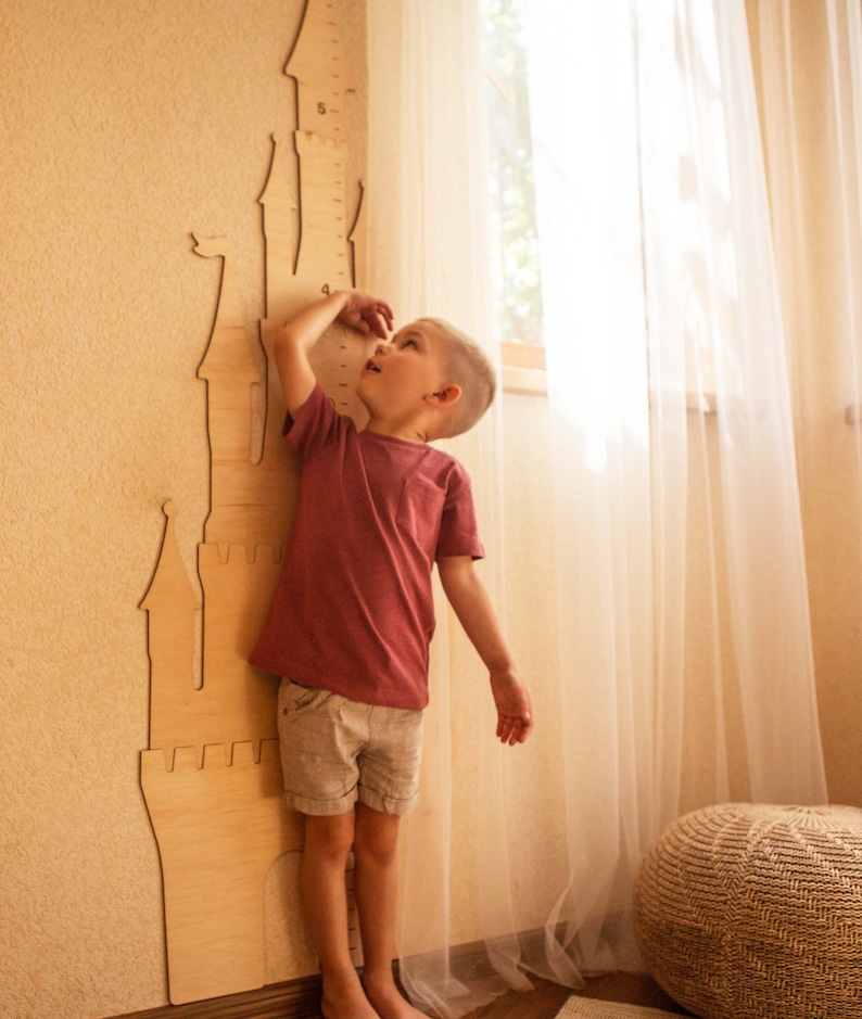 May include: A young child stands next to a wooden castle-shaped height chart, reaching up to check their height. The chart has numbers printed on it, and the child is wearing a pink shirt and shorts.