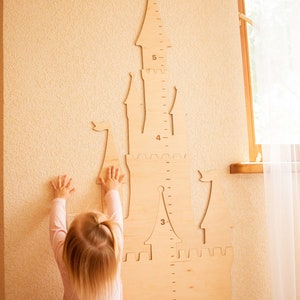 May include: A child stands in front of a wooden castle-shaped height chart. The chart is marked with numbers from 2 to 6. The child is wearing a pink dress and has blonde hair.