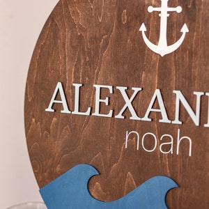 May include: A wooden round sign with a blue and white wave design. The sign has the name "ALEXANDER NOAH" written in white letters and a white anchor.