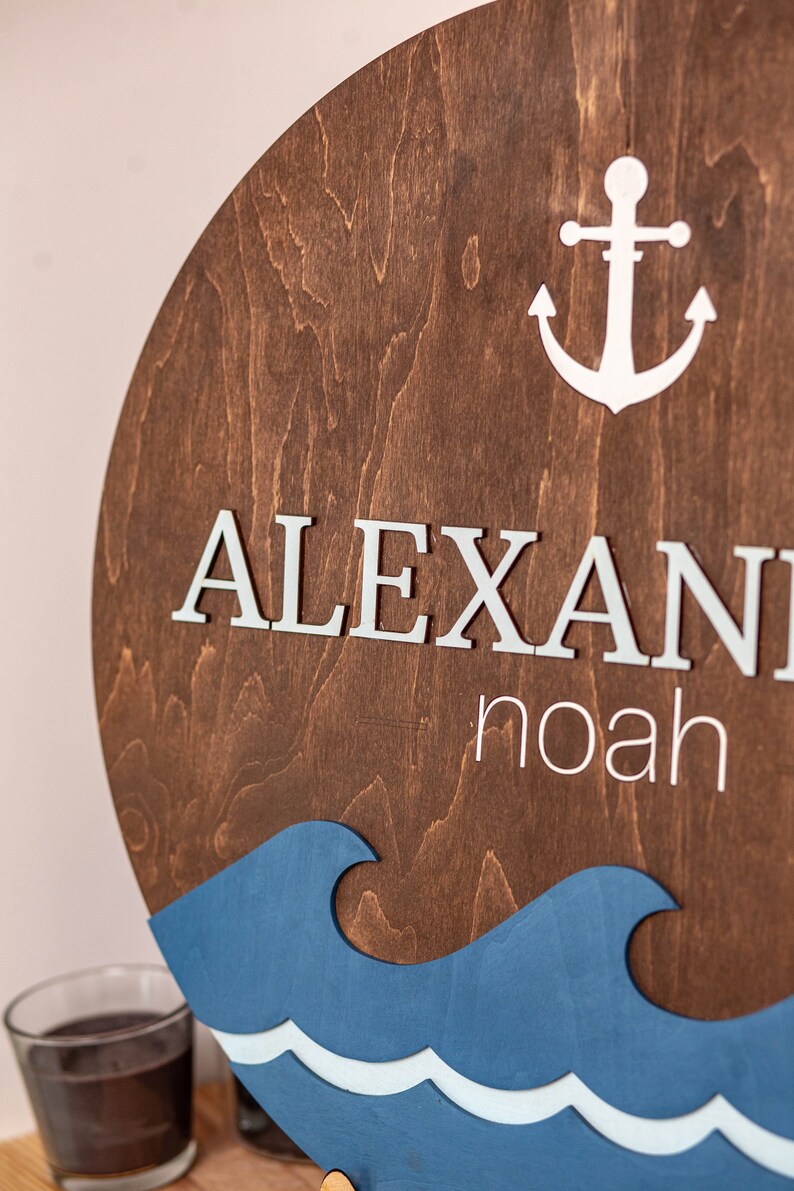 May include: A wooden round sign with a blue and white wave design. The sign has the name "ALEXANDER NOAH" written in white letters and a white anchor.