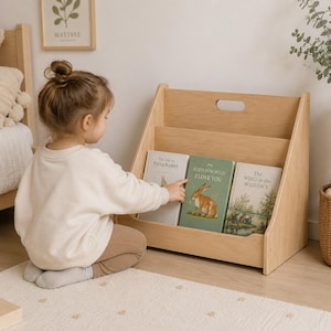May include: A light-colored wooden floor bookshelf with two tiers and a cutout handle at the top. The shelf holds three books titled The Tale of Peter Rabbit, Guess How Much I Love You, and The Wind in the Willows, displayed with their covers facing forward.