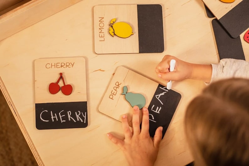 May include: A child is learning to spell fruit names using wooden blocks with chalkboard surfaces. The blocks feature images of a lemon, cherry, and pear. The child is writing the word "pear" on the chalkboard surface of the block.