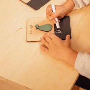 May include: A child's hand writes the letter "P" on a small black chalkboard with white chalk. The chalkboard is next to a wooden block with the word "PEAR" and a green pear shape.