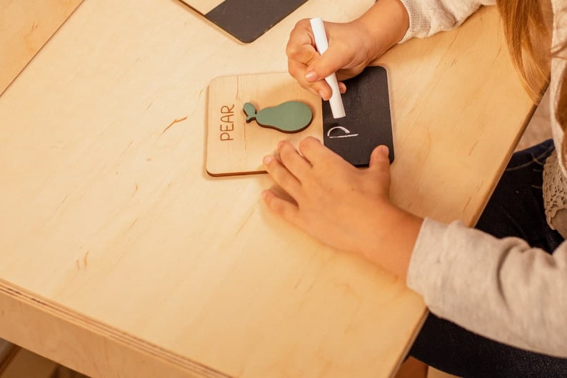 May include: A child's hand writes the letter "P" on a small black chalkboard with white chalk. The chalkboard is next to a wooden block with the word "PEAR" and a green pear shape.