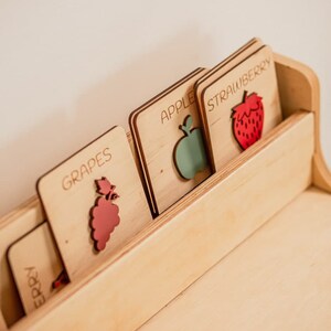 May include: Wooden rack with five cards featuring different fruits. The cards are labeled "Grapes", "Apple", "Strawberry", "Cherry", and "Orange". Each card has a cutout of the fruit.