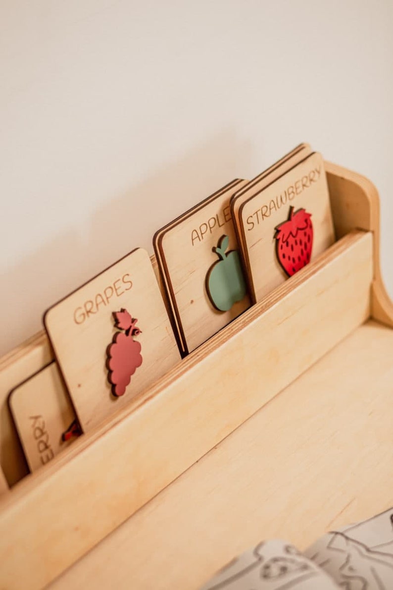 May include: Wooden rack with five cards featuring different fruits. The cards are labeled "Grapes", "Apple", "Strawberry", "Cherry", and "Orange". Each card has a cutout of the fruit.