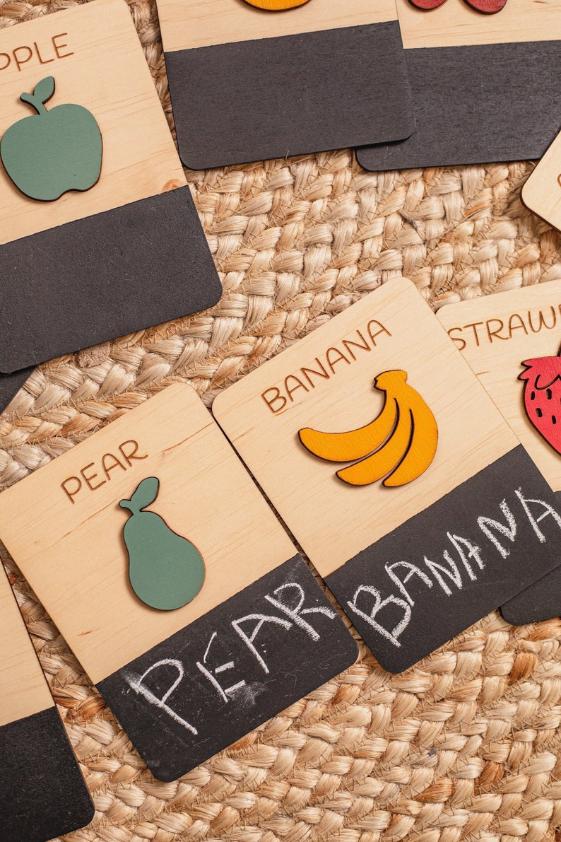 May include: Wooden cards with images of fruits and the names of the fruits written on them. The fruits include an apple, pear, banana, and strawberry. The cards have a black chalkboard surface on the bottom.