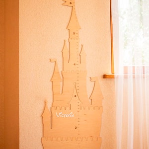 May include: A wooden castle-shaped growth chart with a ruler on the side, measuring from 1 to 6 feet. The castle is painted in a light wood color and has a white inscription that says "Victoria".