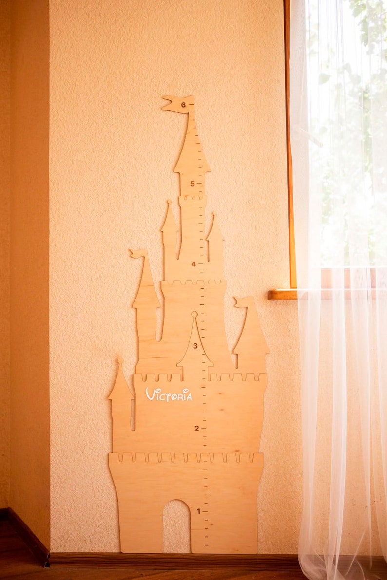 May include: A wooden castle-shaped growth chart with a ruler on the side, measuring from 1 to 6 feet. The castle is painted in a light wood color and has a white inscription that says "Victoria".