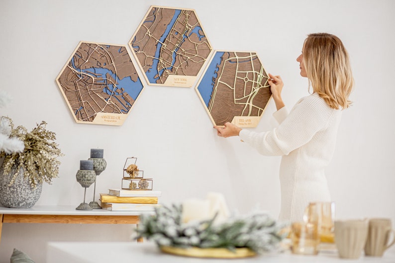 May include: Three hexagonal wooden wall maps with laser-cut city outlines. The maps are in shades of brown, blue, and green. The maps are labeled "Amsterdam", "New York", and "Tel Aviv". A person is hanging the Tel Aviv map on a white wall.