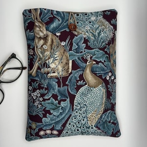May include: A rectangular book sleeve with a button fastening. The fabric displays a pattern of a rabbit, peacock, and foliage in brown, teal, and white on a burgundy backdrop. The button is brown.