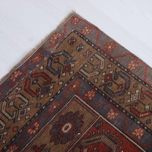 May include: Close-up of a section of a vintage rug with a detailed geometric pattern. The rug features a color palette of brown, red, and blue, with intricate floral and geometric designs. The rug's corner is visible, showing its fringed edge.