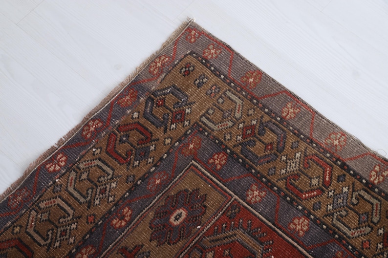 May include: Close-up of a section of a vintage rug with a detailed geometric pattern. The rug features a color palette of brown, red, and blue, with intricate floral and geometric designs. The rug's corner is visible, showing its fringed edge.