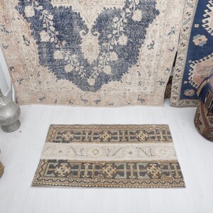 May include: A vintage style rug with a beige background and a dark blue floral pattern. The rug is laid out on a white floor with a smaller rug in front of it. The smaller rug has a beige background with a dark brown and blue geometric pattern.