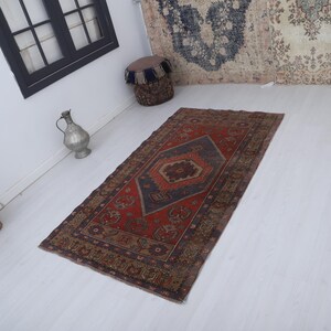 May include: A rectangular, vintage-style rug with a central geometric design in shades of red, navy blue, and brown. The rug features a detailed border and is placed on a white floor, showcasing its intricate patterns.