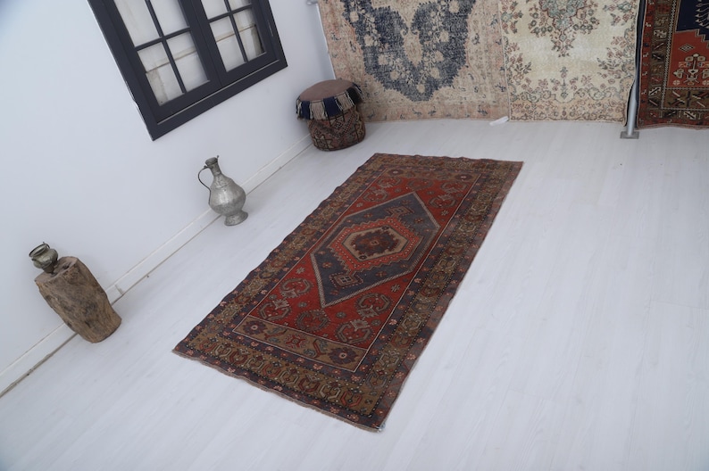 May include: A rectangular, vintage-style rug with a central geometric design in shades of red, navy blue, and brown. The rug features a detailed border and is placed on a white floor, showcasing its intricate patterns.
