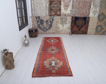 May include: A long, rectangular, red rug with geometric patterns in shades of orange, gray, and blue. The rug is displayed on a light-colored floor, with a window and decorative items in the background. The rug's dimensions are not visible.