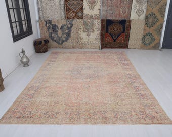 Puede incluir: Una alfombra grande y rectangular de estilo vintage con un aspecto descolorido y desgastado. La alfombra presenta un campo central de tonos rosa y beige apagados, con intrincados patrones florales y geométricos en tonos rojos, azules y crema. La alfombra se exhibe sobre un suelo de color claro.
