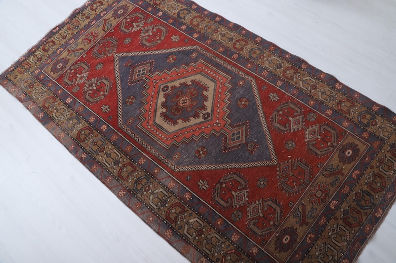 May include: A rectangular, vintage-style rug with a central diamond pattern in shades of red, blue, and brown. The rug features intricate geometric designs and a distressed appearance, suggesting it is an antique or a well-loved piece.