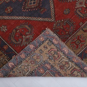 May include: A section of a vintage rug with a geometric pattern. The rug features a central red field with dark blue and tan borders. The design includes repeating shapes and floral motifs.