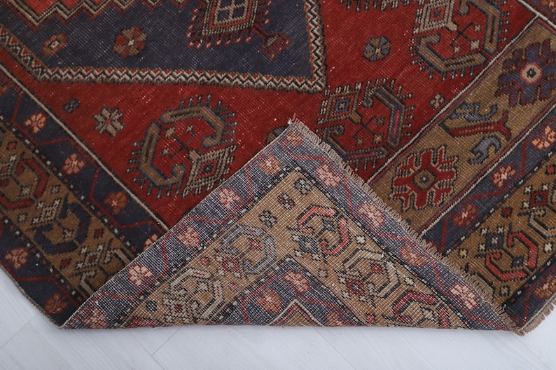 May include: A section of a vintage rug with a geometric pattern. The rug features a central red field with dark blue and tan borders. The design includes repeating shapes and floral motifs.