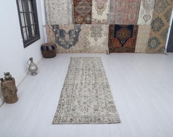 Puede incluir: Una alfombra rectangular de color beige con un patrón oscuro y detallado. La alfombra está colocada sobre un suelo blanco, con otras alfombras colgadas en el fondo. El diseño de la alfombra presenta elementos florales y geométricos.