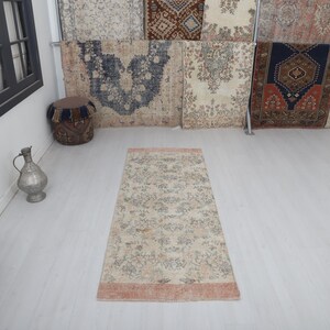 May include: A rectangular rug with a floral pattern and a peach border. The rug features a cream background with green and blue floral designs. It is placed on a light-coloured wooden floor. Various antique rugs are displayed in the background.