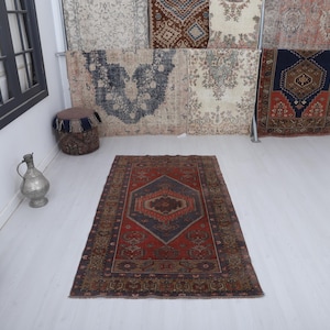 May include: A rectangular, vintage rug with a central diamond pattern in shades of red, blue, and brown. The rug has a detailed border and is displayed on a white floor. Decorative items are in the background.