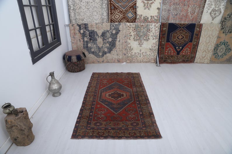 May include: A rectangular, vintage rug with a central diamond pattern in shades of red, blue, and brown. The rug has a detailed border and is displayed on a white floor. Decorative items are in the background.