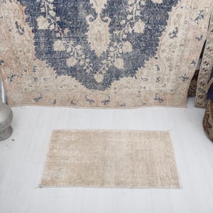 May include: A small, beige rug with a distressed, vintage look. The rug is laid out on a white surface.