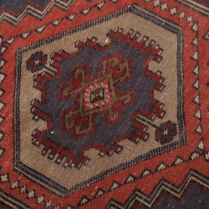May include: Close-up of a section of a vintage rug with a geometric pattern. The rug features a central medallion in shades of blue, brown, and red, surrounded by a red border with zigzag designs. The rug has a worn, textured appearance.