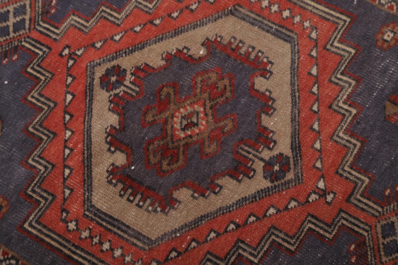 May include: Close-up of a section of a vintage rug with a geometric pattern. The rug features a central medallion in shades of blue, brown, and red, surrounded by a red border with zigzag designs. The rug has a worn, textured appearance.