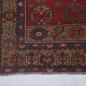 May include: Close-up of a section of a vintage rug with a detailed geometric pattern. The rug features a rich color palette of red, brown, and navy blue. The design includes repeating floral and geometric motifs, with a fringed edge.