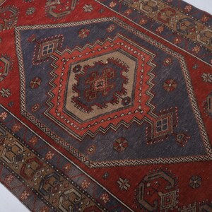 May include: A rectangular rug with a central diamond pattern in shades of red, blue, and brown. The rug features intricate geometric designs and a border with repeating motifs. The rug's color palette and patterns suggest a traditional or vintage style.