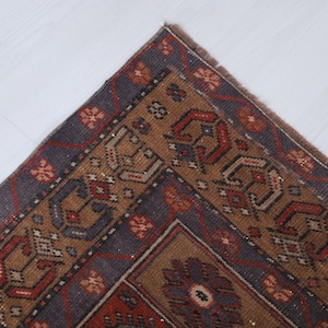May include: A section of a vintage rug with a detailed geometric pattern. The rug features a central design in shades of brown, red, and blue, with a dark blue border. The corner of the rug is visible, showcasing the intricate craftsmanship.