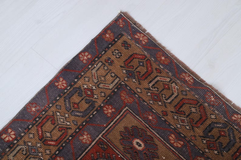 May include: A section of a vintage rug with a detailed geometric pattern. The rug features a central design in shades of brown, red, and blue, with a dark blue border. The corner of the rug is visible, showcasing the intricate craftsmanship.