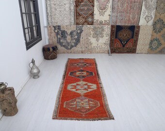 May include: An orange and beige runner rug with geometric patterns. The rug features a central design with diamond and star-like shapes in shades of blue, beige, and orange. The rug is displayed on a light-colored floor, with a collection of rugs hanging in the background.