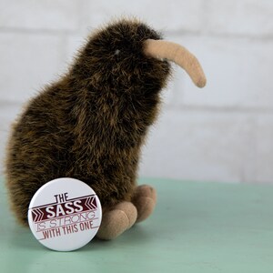 May include: A brown and white stuffed kiwi bird with a white button that says "The Sass is Strong with This One" on a light green surface.