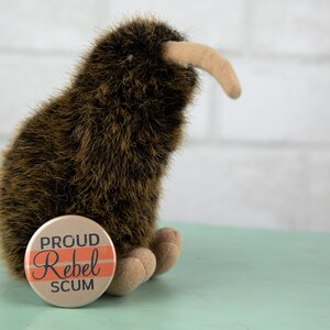 May include: A brown and black stuffed kiwi bird with a long beak. The kiwi is sitting on a light green surface with a round button that says "Proud Rebel Scum" in black text with orange stripes.