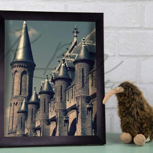 May include: A framed photograph of a medieval-style building with multiple towers. The building is made of brick and stone and has a dark, aged appearance. The photo is in a black frame and is hanging on a white brick wall.