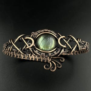 May include: A wire-wrapped cuff bracelet featuring a central round labradorite gemstone with iridescent green and gray tones. The bracelet is crafted from oxidized copper wire, showcasing intricate woven patterns and decorative swirls.