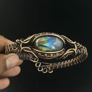 May include: A wire-wrapped cuff bracelet featuring a central oval labradorite gemstone with blue and gold iridescence. The bracelet is constructed from dark oxidized copper wire, intricately coiled and woven into a decorative frame.