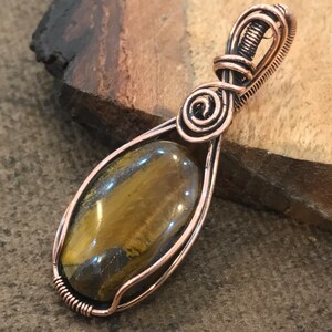 May include: A teardrop-shaped tiger's eye gemstone pendant encased in a decorative copper wire wrap. The wire features intricate coils and a spiral design at the top, forming a bail for a necklace chain. The stone displays brown and gold tones.
