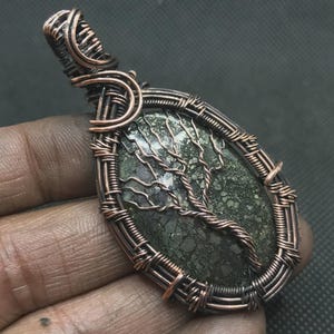 Può includere: Ciondolo avvolto in filo di rame con un centro in pietra verde ovale. La pietra presenta un design dettagliato ad albero in filo di rame. Il ciondolo è tenuto in mano su uno sfondo scuro. Il filo ha un colore rame naturale invecchiato.