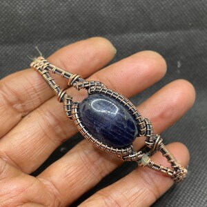 Op de afbeelding: Een handgemaakte, met draad omwikkelde hanger met een donkerblauwe, ovaalvormige edelsteen. De koperdraad is ingewikkeld rond de steen geweven en vormt een rechthoekig frame. De hanger wordt in een hand gehouden.