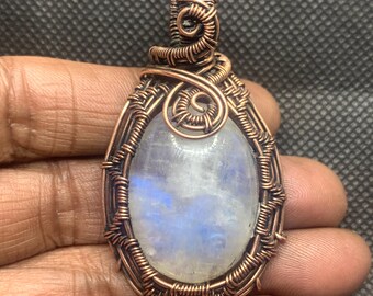 Peut inclure: Un pendentif en pierre de lune de forme ovale enchâssé dans un fil de cuivre complexe enroulé à la main. La pierre précieuse présente un aspect blanc translucide avec une adularescence bleue. Le fil de cuivre forme un cadre décoratif.