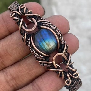 May include: A wire-wrapped bracelet featuring a central oval labradorite gemstone with blue and gold iridescence. The stone is encased in intricate, dark-toned copper wire coils and loops that form a decorative, textured band structure.